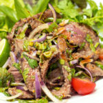 A plate of Budget-Friendly Grilled Beef Salad with Homemade Thai Dressing, featuring mixed greens, sliced tomatoes, green chili peppers, and red onion, topped with grilled flank steak. This Weight Watchers-friendly salad is packed with flavor and nutrition, making it a perfect option for those following the WW Plan