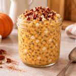 Jar of pumpkin pie cottage cheese overnight oats with chia seeds, topped with chopped pecans and cinnamon, with a pumpkin and a bowl of pecans beside it